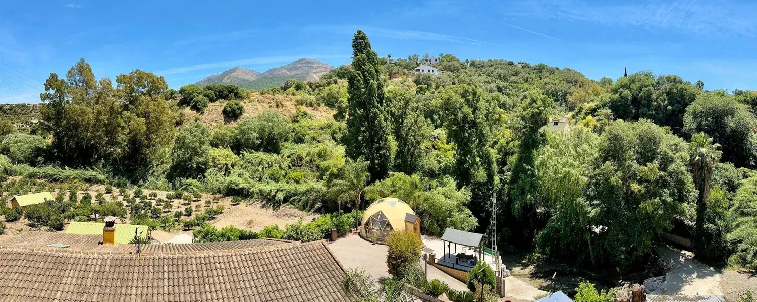 Scenic view of a lush, green hillside with trees, houses, and mountains in the background on a bright, sunny day - retreat center of From gaia for you