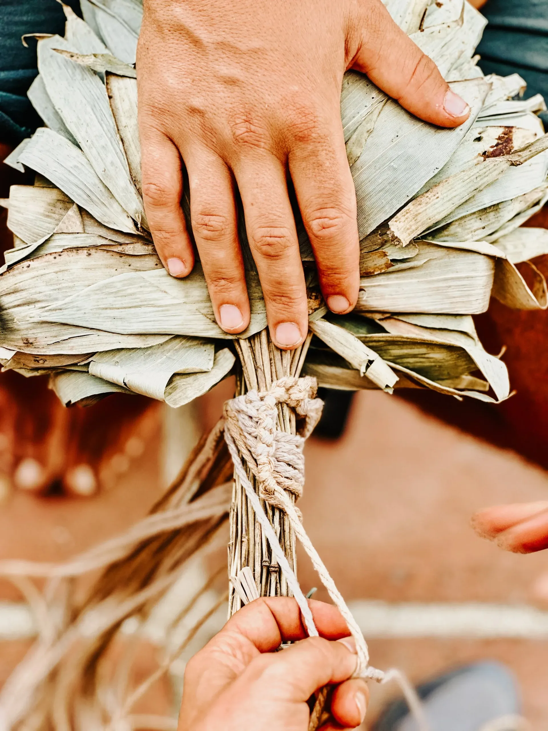 Close-up of a person's hand holding a bundle of dried tropical leaves wrapped with string, with another hand reaching in.