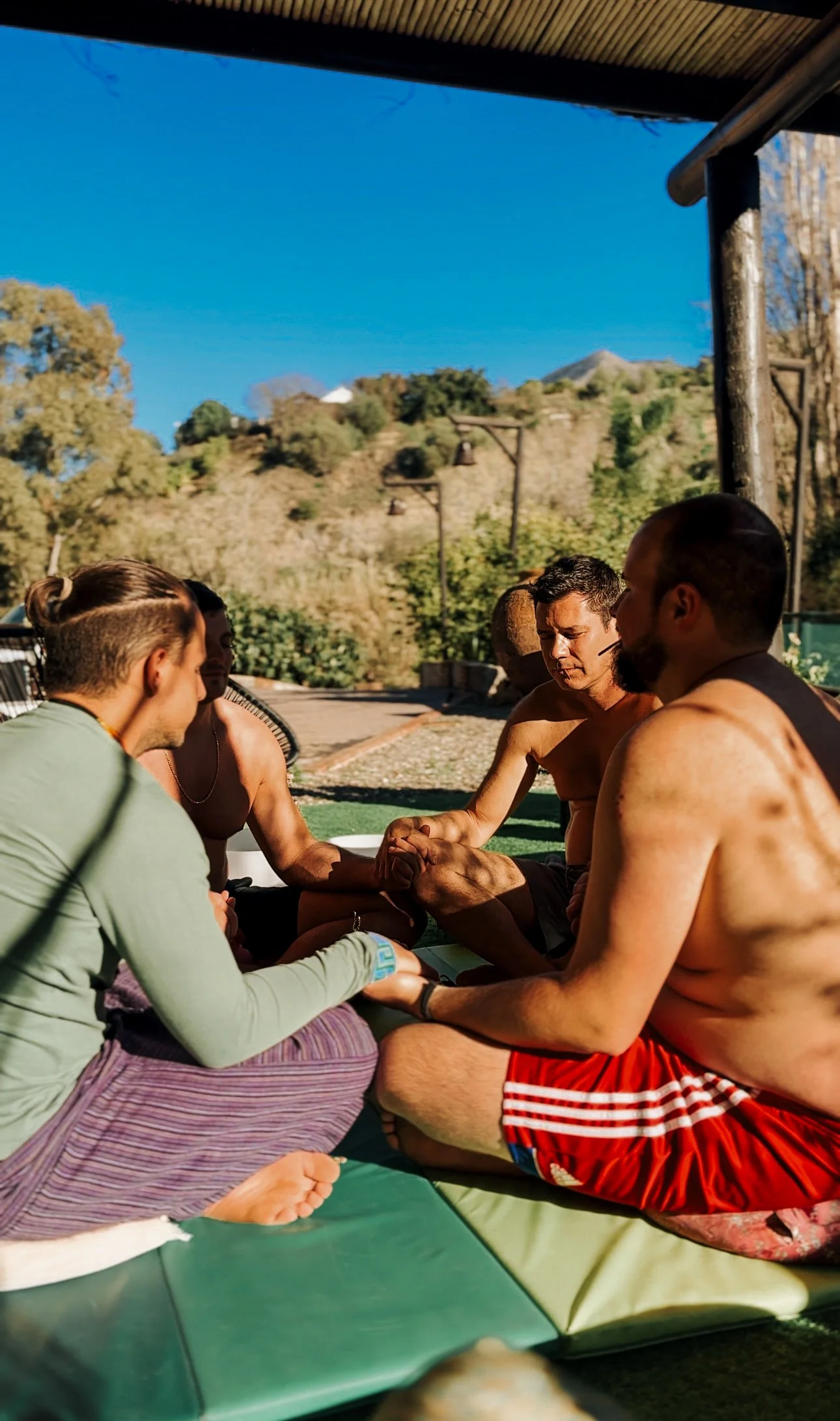 Group of men sitting outdoors, some shirtless, engaging in a handshake or discussion, with a scenic hillside and blue sky in the background.